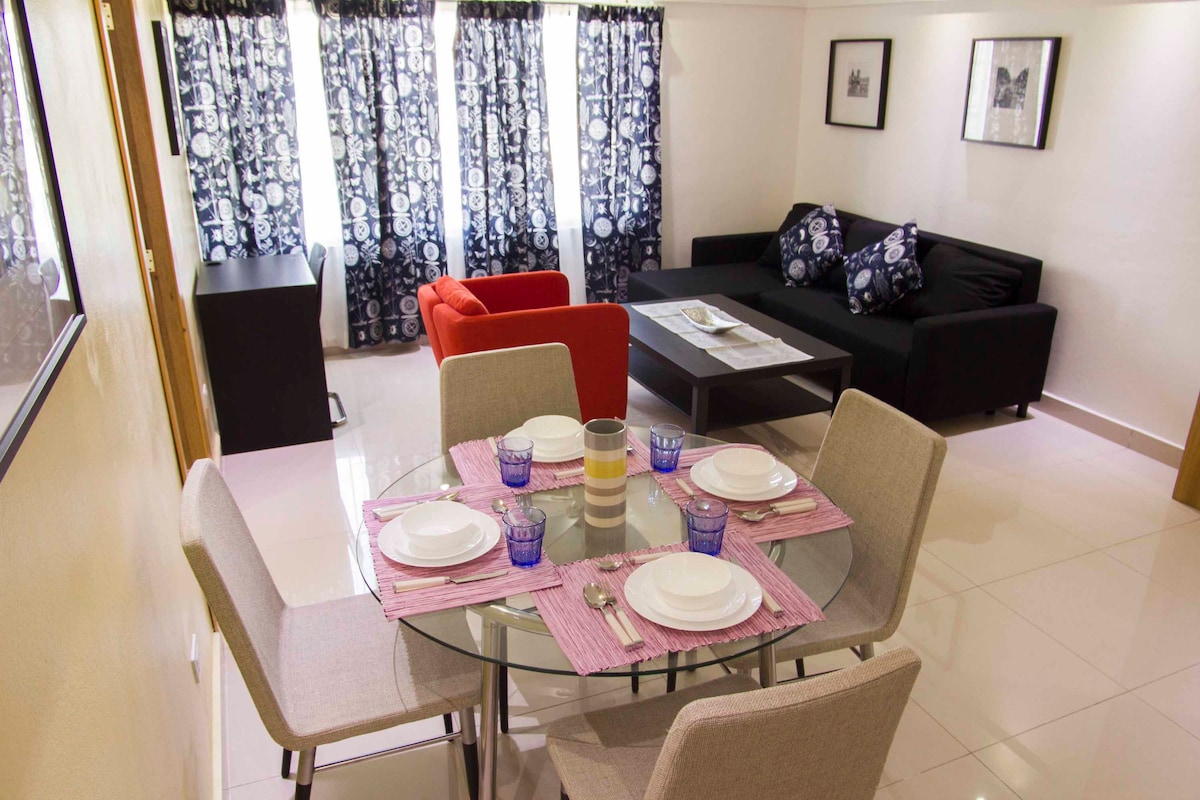 A modern dining area showcases a round table set with plates and utensils, surrounded by upholstered chairs. A cozy living space features a black couch with decorative pillows and a vibrant red chair, with large windows draped in patterned curtains allowing natural light.