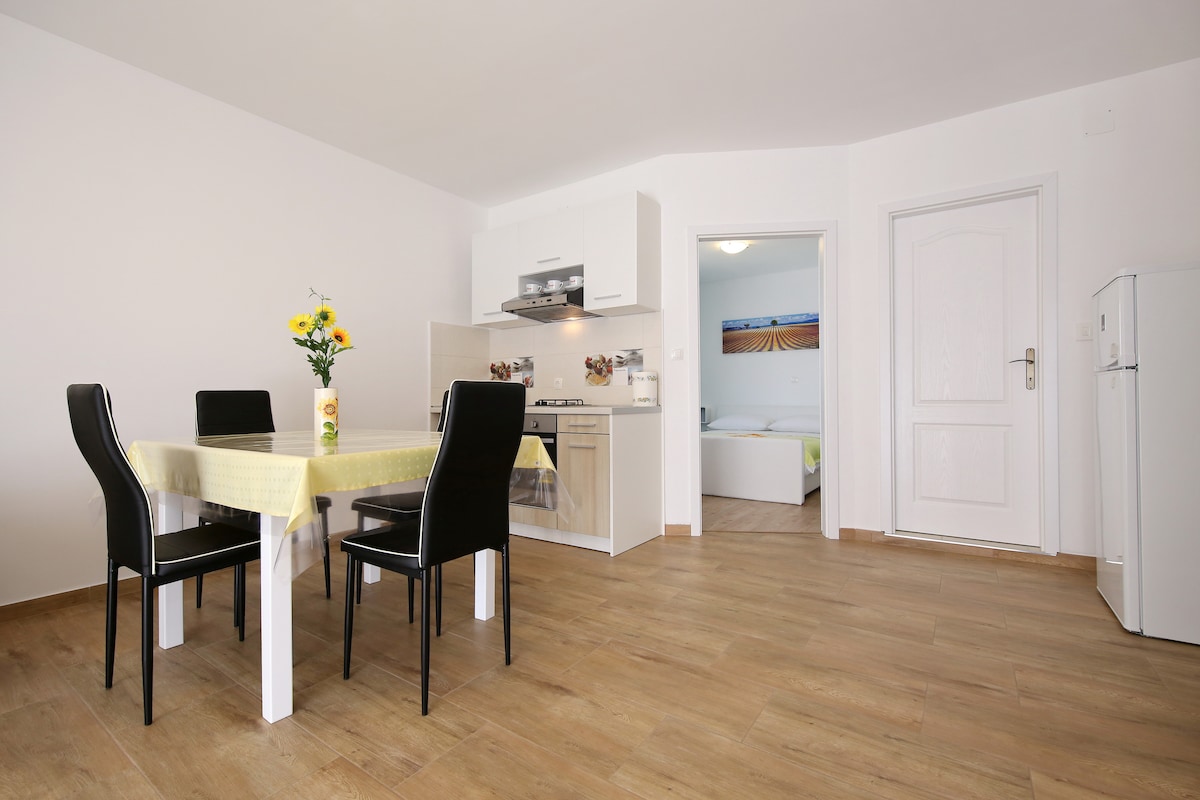 A modern kitchen and dining area are visible, featuring a round table set with four black chairs. A gas stove and oven are situated against the wall, accompanied by a refrigerator. A vase of sunflowers adds a touch of color to the table, while light wood flooring enhances the space.