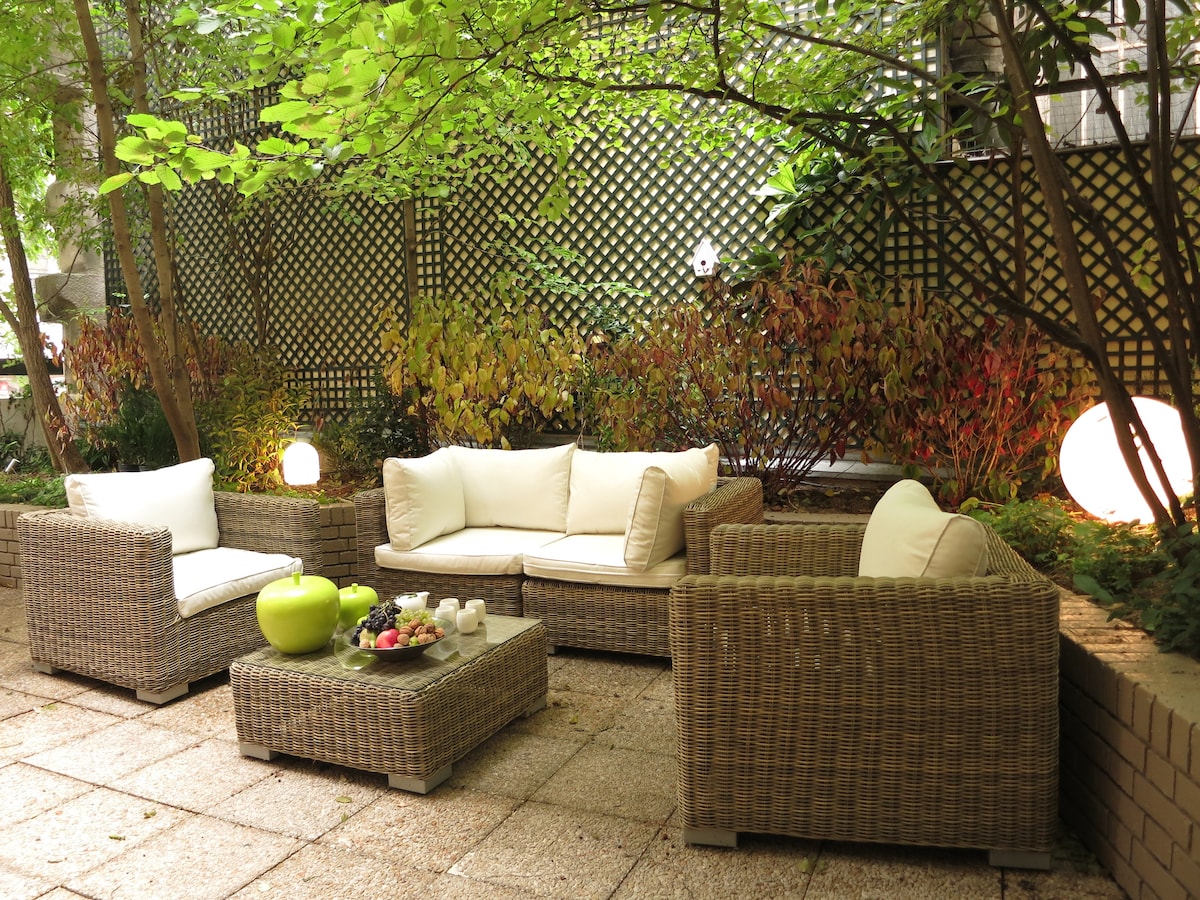 A tranquil garden terrace is furnished with a seating area featuring wrapped wicker furniture and soft cream cushions. A low coffee table with decorative items, including green apples and small candles, complements the surrounding foliage and vibrant plants.