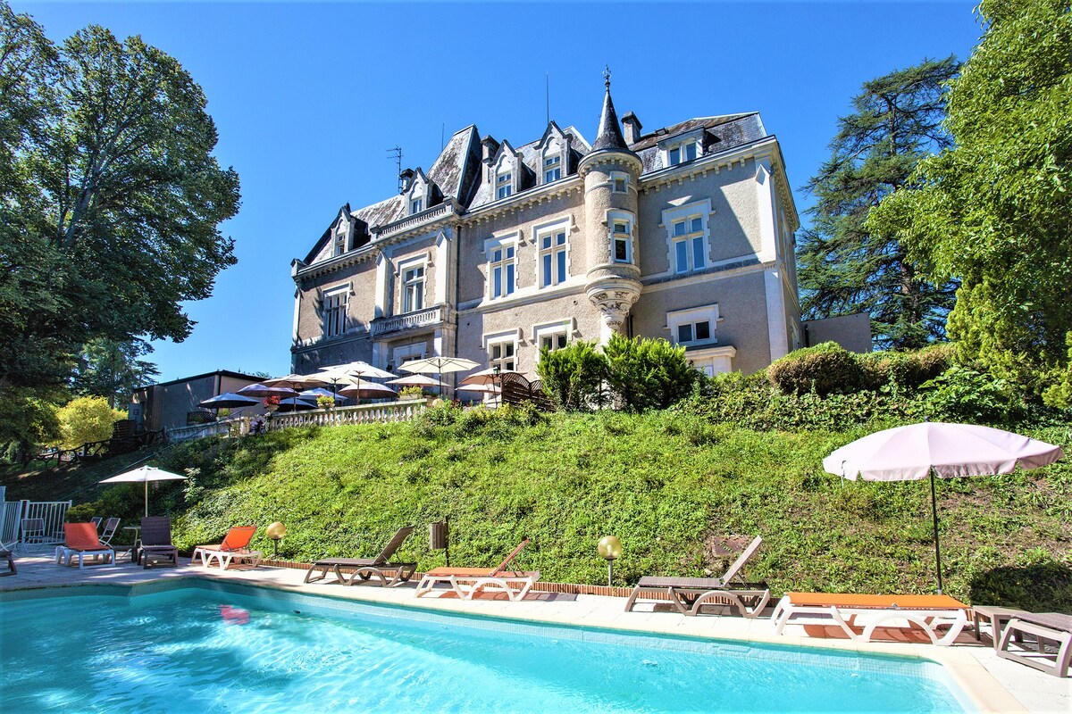 The historic Château des Reynats is depicted against a clear blue sky, showcasing its elegant architecture. A swimming pool lies in the foreground, surrounded by sun loungers and umbrellas. Lush greenery enhances the tranquil atmosphere, inviting relaxation and leisure.