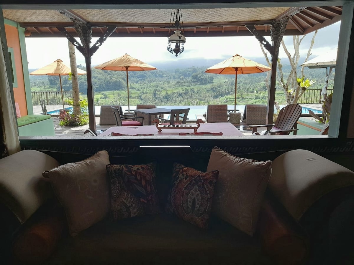 A cozy seating area is framed by a large window, offering a view of the outdoor terrace furnished with sun umbrellas and lounge chairs. Lush greenery and distant hills are visible beyond, revealing the serene landscape of Bali.