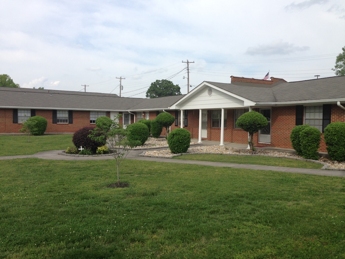 A well-maintained exterior of a single-story apartment complex is showcased, surrounded by neatly manicured lawns and shrubs. Pathways lead through the landscaping, providing access to the individual units. A connection to the quiet neighborhood is indicated by the low-rise brick structure.