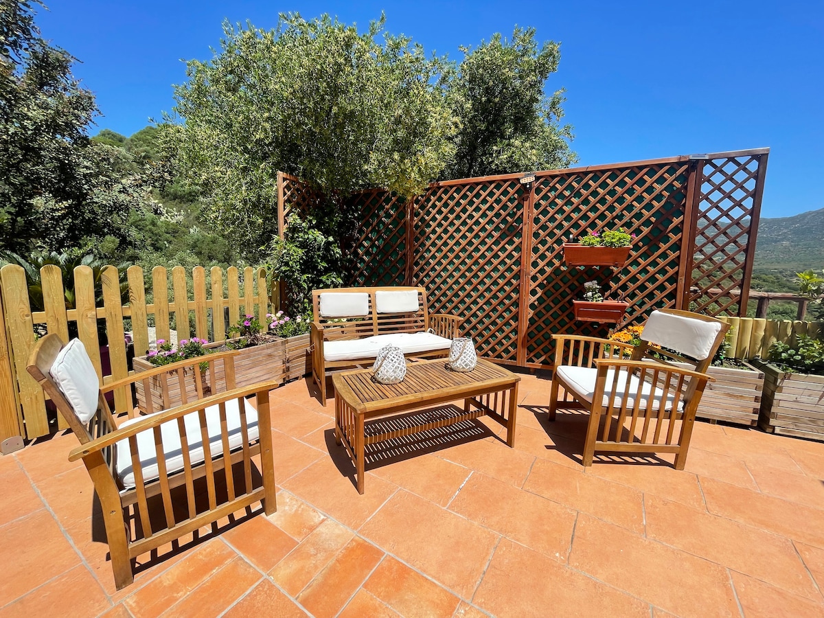 A spacious outdoor seating area features wooden furniture, including a coffee table surrounded by four cushioned chairs. Greenery and flowers provide a natural backdrop, with a wooden lattice offering privacy. The sunlit terrace highlights the warm tones of the terracotta flooring.