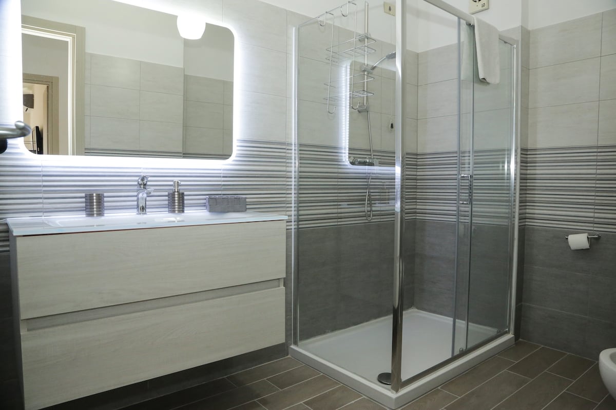 A spacious bathroom is featured, showcasing a modern shower with glass doors and a sleek vanity with dual sinks. Soft lighting is provided by a backlit mirror, highlighting the neutral tones of the tiled walls and floor.