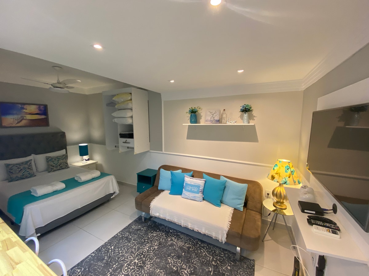 A well-organized studio space features a spacious bed with white linens and complementary blue accents. A brown sofa with decorative pillows is positioned nearby, and a wall-mounted TV is visible. Soft lighting is provided by a stylish lamp on a side table, enhancing the inviting ambiance.