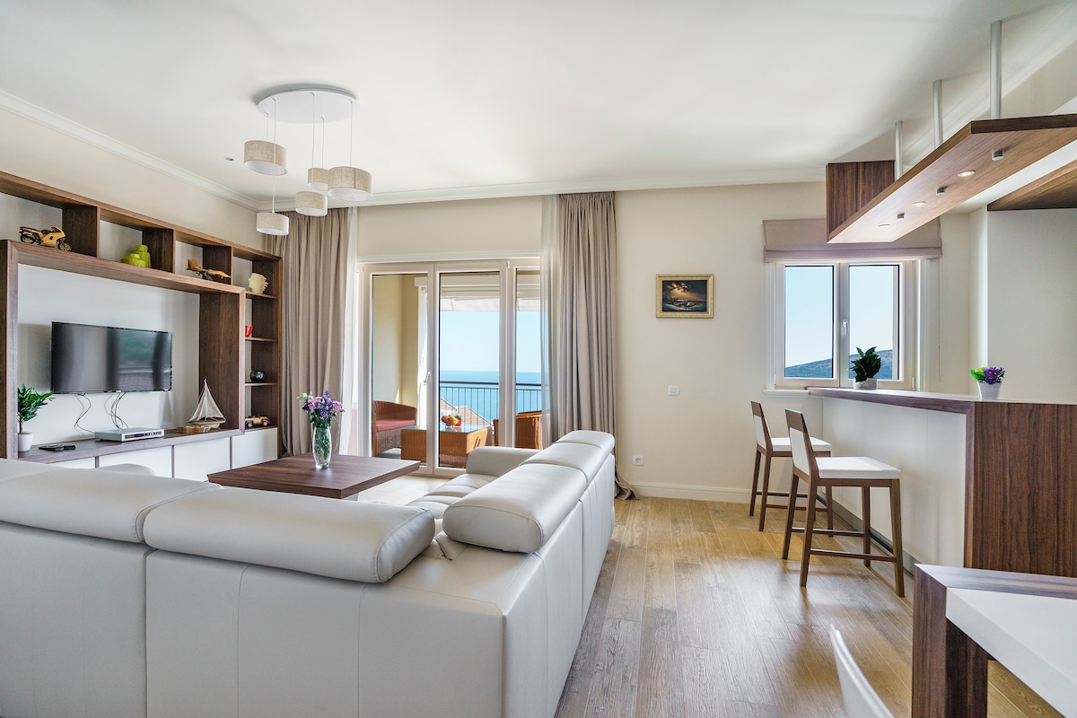 A bright living area features a white sofa and a wall-mounted TV. Large windows provide a view of the sea. The open concept design includes a kitchen bar with stools, allowing for easy interaction between spaces.
