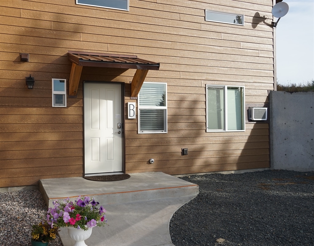 The exterior of the duplex showcases a brown wooden façade with crisp white trim. A covered entry features a white door marked with a letter 'B'. Nearby, a decorative pot filled with colorful flowers adds a welcoming touch to the pathway.