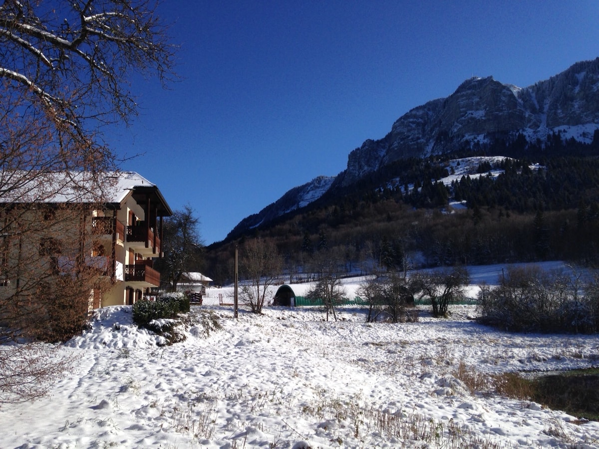 A chalet-style building is seen with snow-covered surroundings, highlighting its balconies and the natural landscape. Mountains rise majestically in the background, providing a scenic backdrop. The ground is blanketed in fresh snow, while trees and bushes create a tranquil atmosphere.