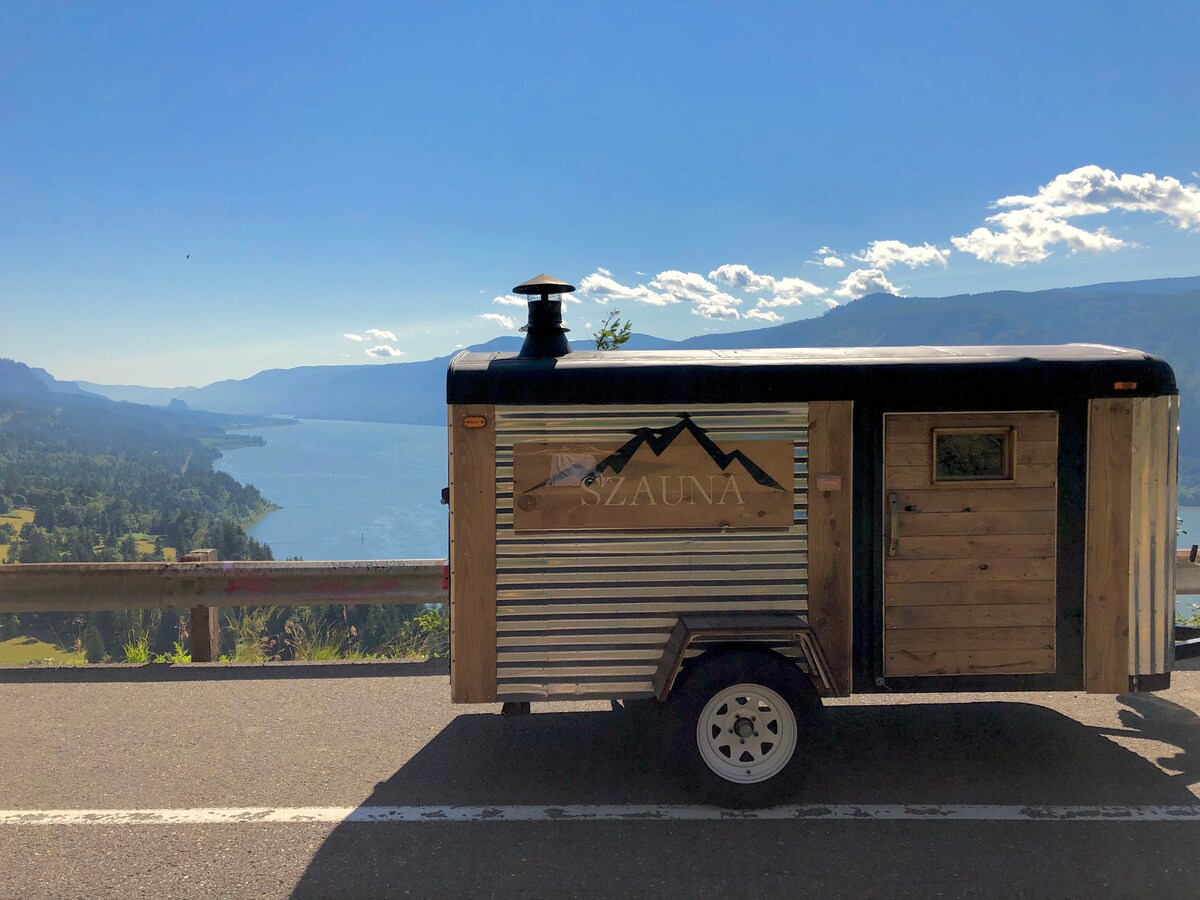 A mobile sauna trailer is positioned along a scenic roadside, overlooking a vast body of water and mountainous terrain. The exterior features a combination of wood and metal materials, with a chimney visible at the top. Clear skies and fluffy clouds enhance the natural landscape.