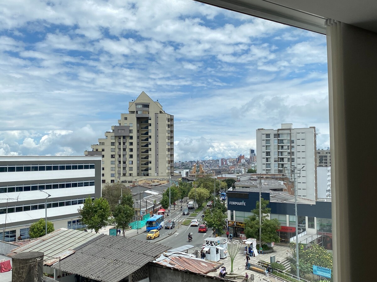 A view of the city is captured through a large window, showcasing a mix of modern buildings and local architecture. The sky is partly cloudy, and several vehicles are seen on the street. Trees and shops are visible, adding to the urban landscape.