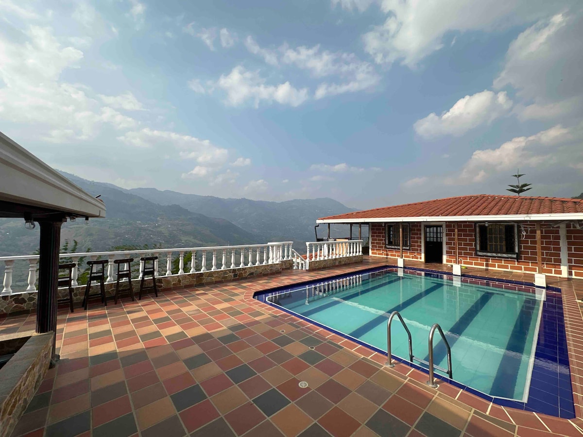 A large outdoor swimming pool is bordered by colorful tiled flooring. The space features a scenic backdrop of rolling hills and mountains, with a spacious terrace that includes seating areas. The building showcases a rustic brick facade, adding character to the tranquil environment.