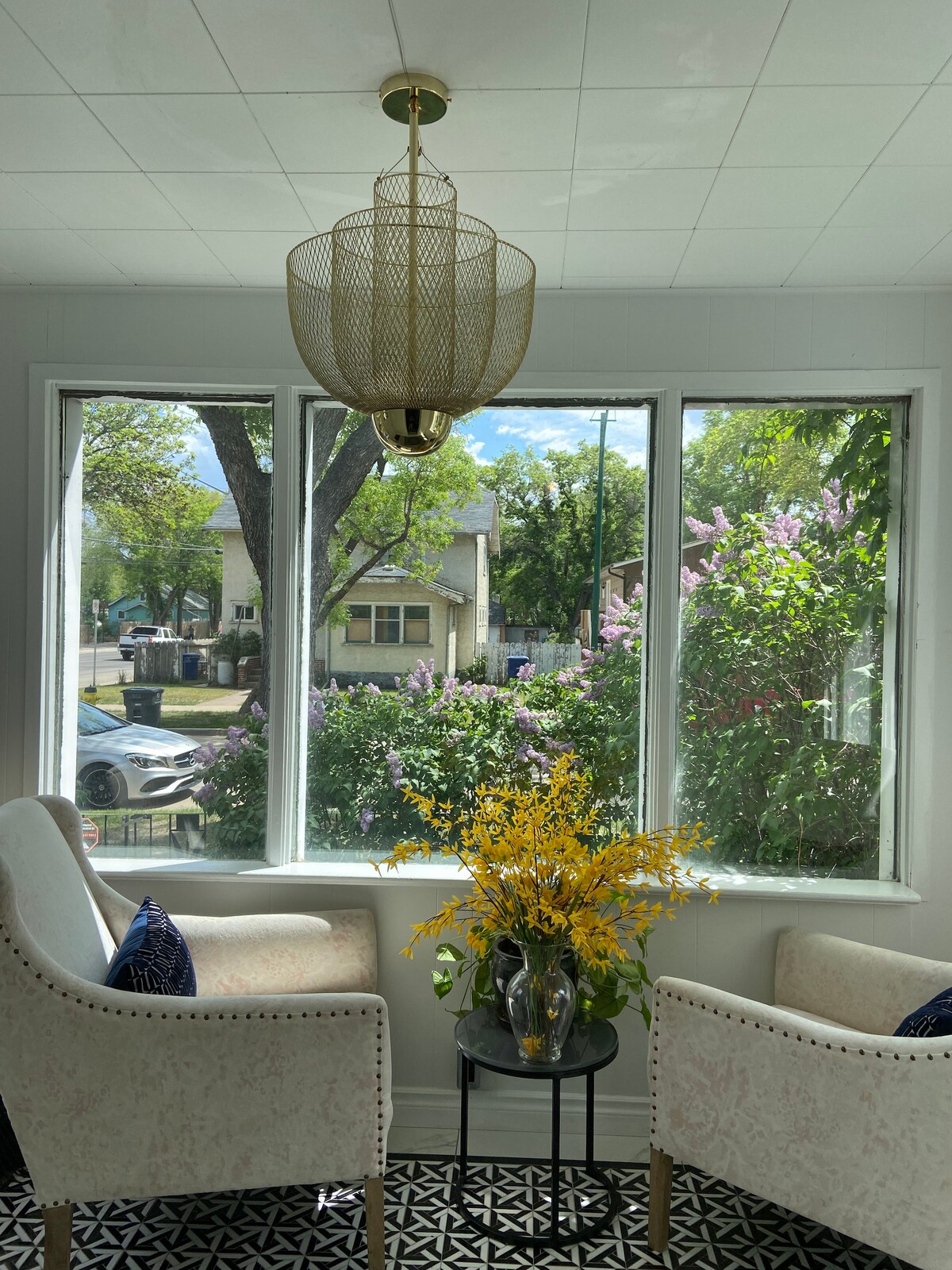 A bright and inviting sitting area features two chairs positioned by large windows. Fresh greenery and blooming flowers are visible outside, creating a vibrant backdrop. A small table holds a decorative vase filled with yellow flowers, adding a pop of color to the space.