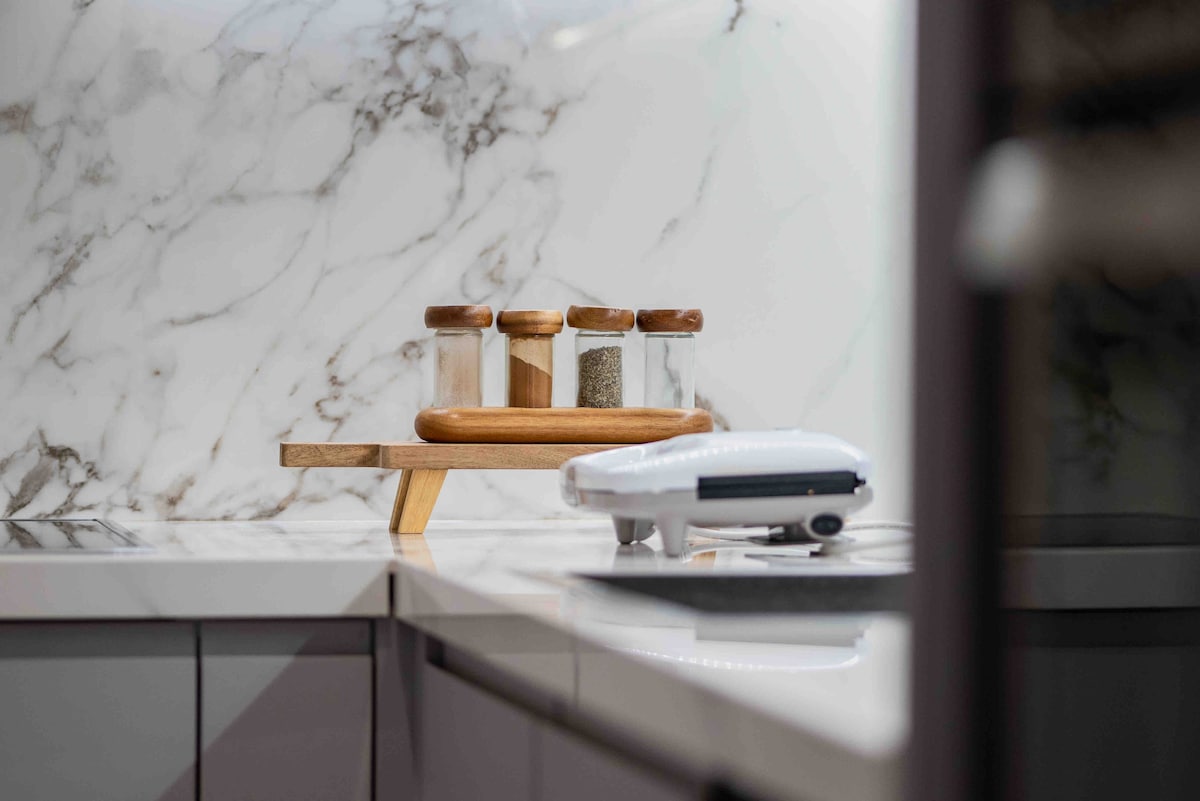 A modern kitchen countertop features a marble backsplash, showcasing a wooden spice rack with glass jars filled with various spices. A sleek kitchen appliance is positioned nearby, highlighting the functionality of the space.
