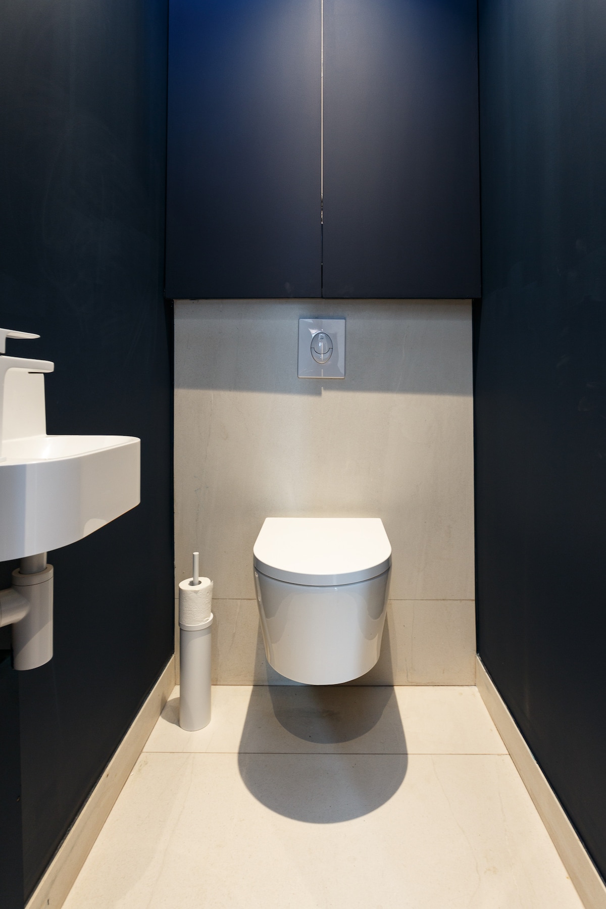 The image showcases a compact bathroom space featuring a modern toilet and a minimalist sink. A sleek white toilet is positioned against a navy blue wall, with a small cabinet above for storage. A cylindrical toilet brush is neatly placed beside the toilet.