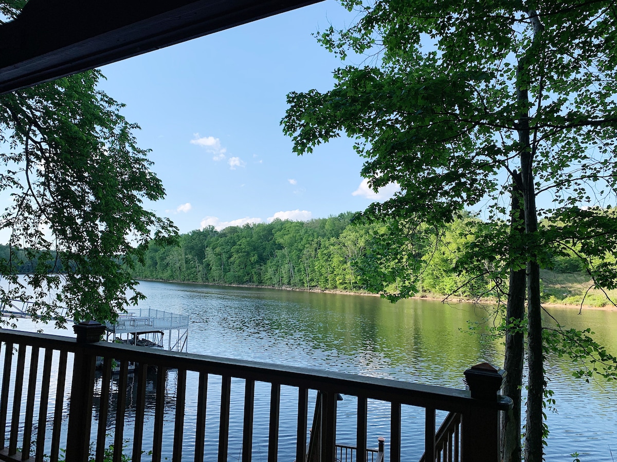 A calming view of the river is framed by lush green trees. The water's surface reflects the sky and surrounding foliage, creating a serene atmosphere. A wooden railing is visible in the foreground, adding a natural element to the scenic vista.