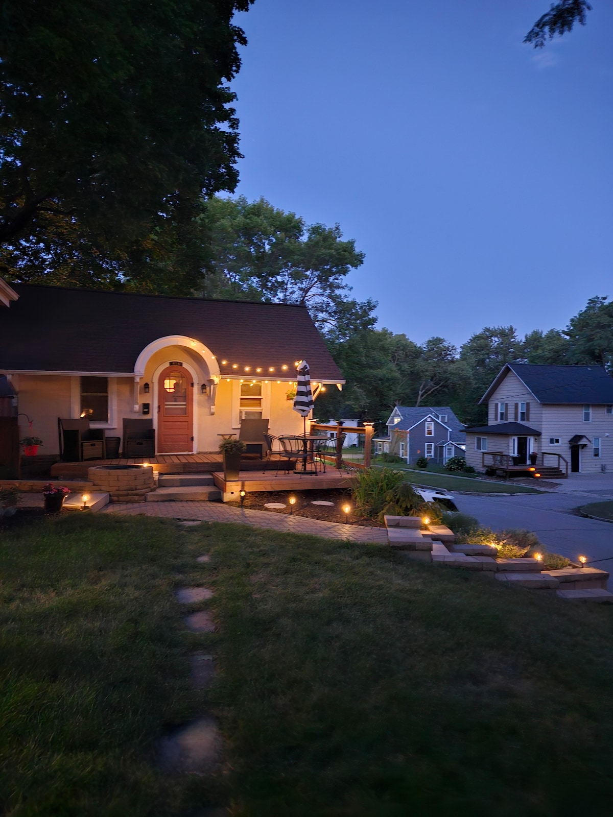 The charming cottage is viewed from a distance, featuring a well-lit entrance with string lights and a landscaped pathway. The inviting porch is furnished with chairs, and a soft glow from the surrounding gardens enhances the tranquil evening ambiance.