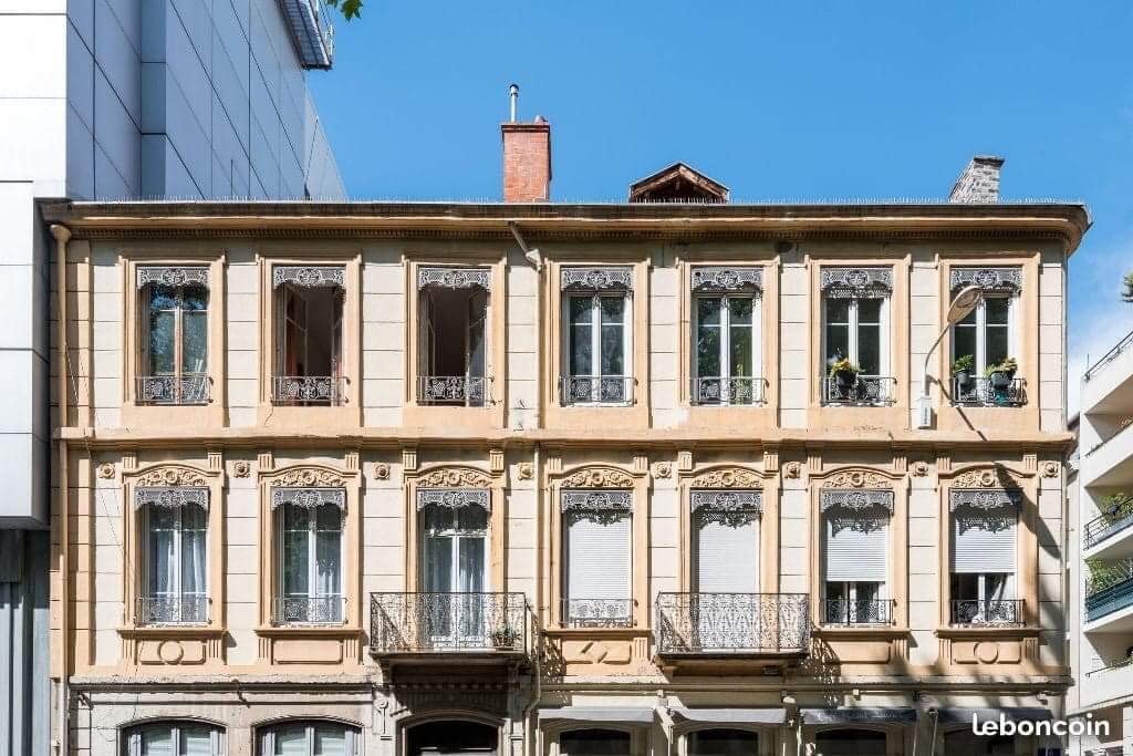The historical building features a classic façade with intricate architectural details. Several windows are adorned with decorative wrought iron balconies, providing charm and character. The structure is surrounded by greenery, contributing to a pleasant streetscape.
