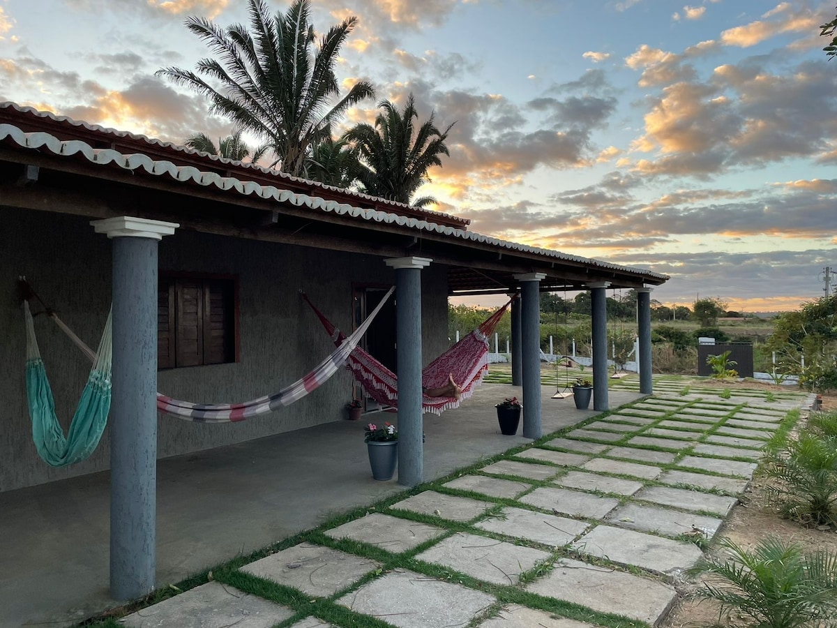 A shaded porch area features several hammocks suspended between columns. The ground is paved with stone tiles arranged in a grid pattern, leading to planted greenery. The backdrop showcases a colorful sky at dusk with scattered clouds and silhouettes of palm trees.