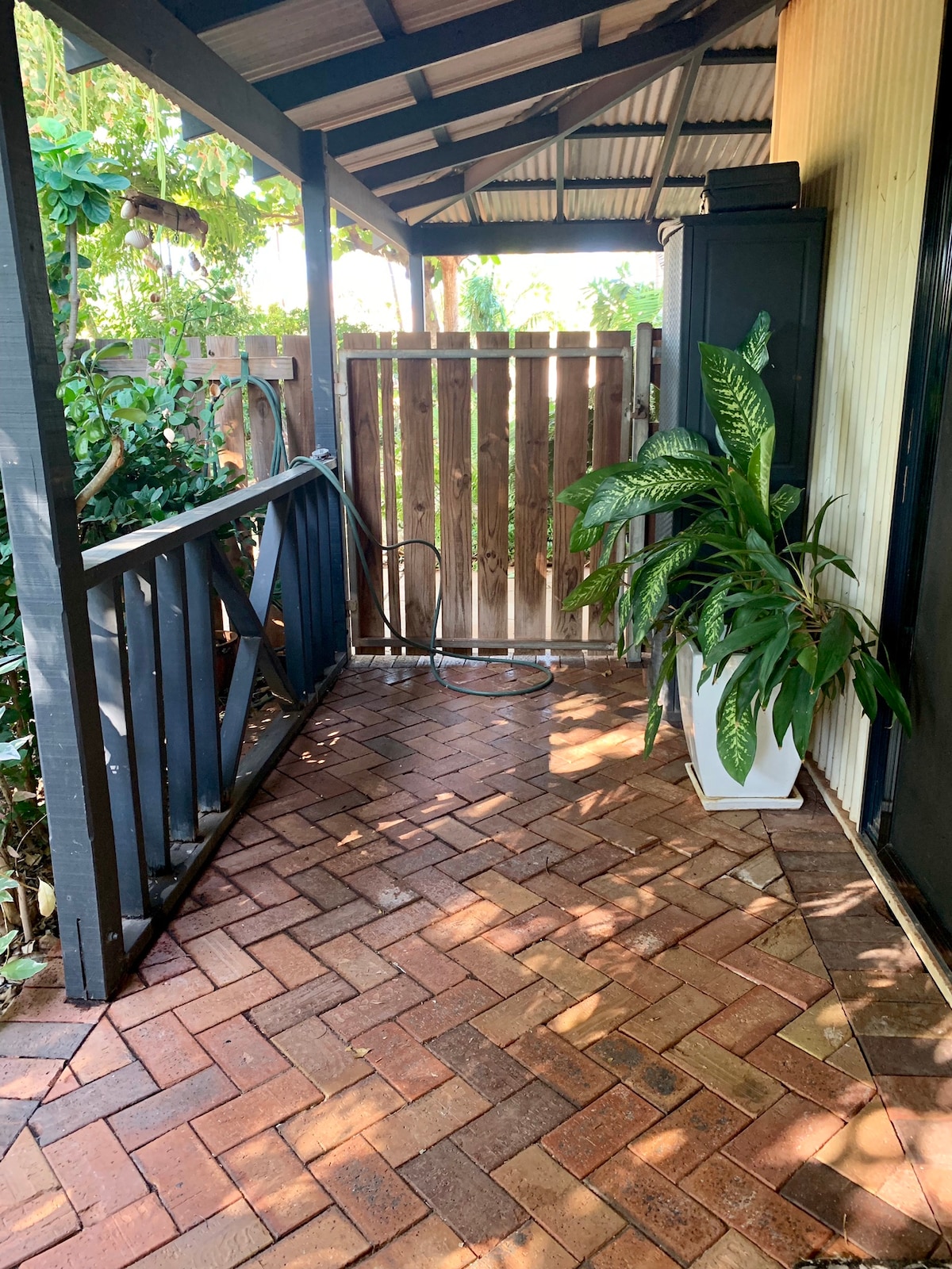 A sheltered entryway features a paved brick pathway leading to a wooden gate. Lush greenery is visible beside the pathway, with a tall potted plant placed against a wall. The area is well-lit and invites visitors to explore further.