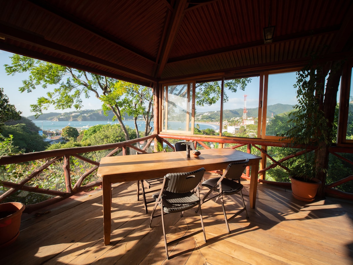 A spacious deck area is surrounded by large windows, offering views of the bay and lush greenery. A wooden table with chairs is positioned in the center, complemented by potted plants that enhance the natural ambiance.