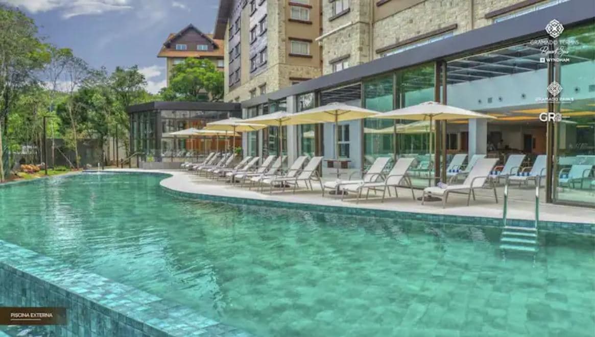 A serene swimming pool is surrounded by lounge chairs arranged under umbrellas, inviting relaxation. The pool's clear turquoise water reflects the greenery and modern architecture of the adjoining buildings, enhancing the tranquil ambiance of the outdoor area.