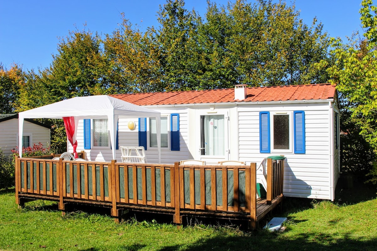 A mobile home is situated within a grassy area, featuring a wooden terrace surrounded by trees. A white awning provides shade over part of the deck, which includes seating. Bright blue shutters frame the windows, complementing the white exterior of the home.