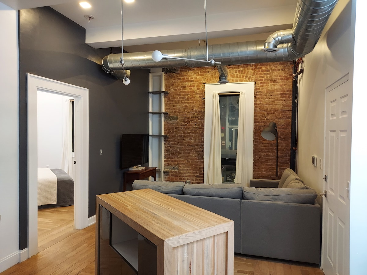 A spacious living area is depicted with a large sectional couch facing a wall-mounted TV. Exposed brick adds texture to the space, complemented by a wooden island in the foreground. Natural light enters through large windows, illuminated by a contemporary lamp.