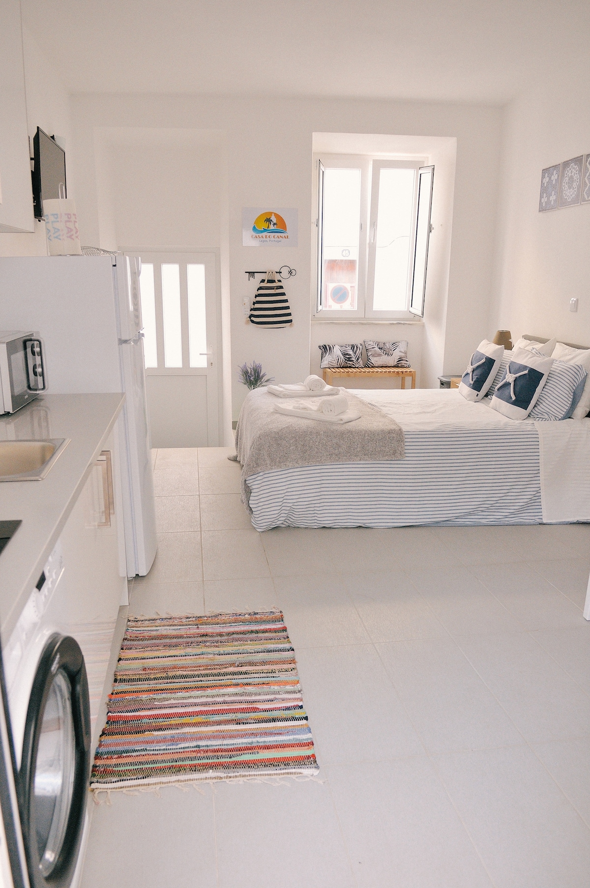 A modern studio space features a cozy sleeping area with a spacious bed dressed in striped linens. A small kitchen with essential appliances, including a refrigerator and washing machine, is visible. Natural light filters in through the two windows, enhancing the airy atmosphere.