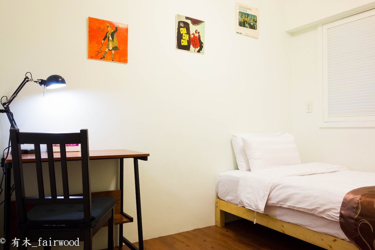 A small room is featured, equipped with a spacious bed and a desk area. A chair is positioned at the desk, illuminated by a desk lamp. Colorful wall art adds character above the bed. A window allows natural light to enter the room.