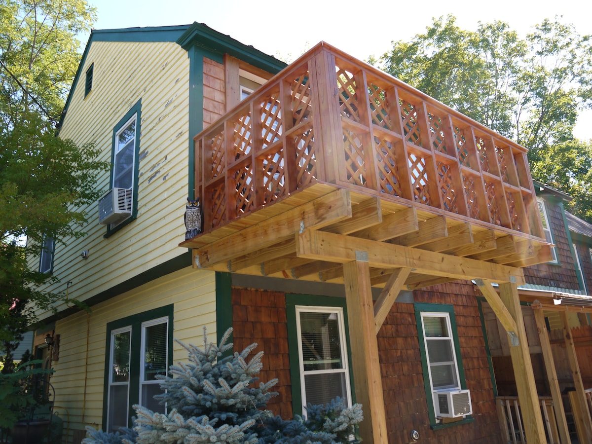 The exterior of a yellow and wooden house is depicted, showcasing a deck supported by a sturdy framework. Lush greenery surrounds the property, highlighting the inviting features of the home. Air conditioning units are visible on the side of the house, suggesting comfort during warmer weather.