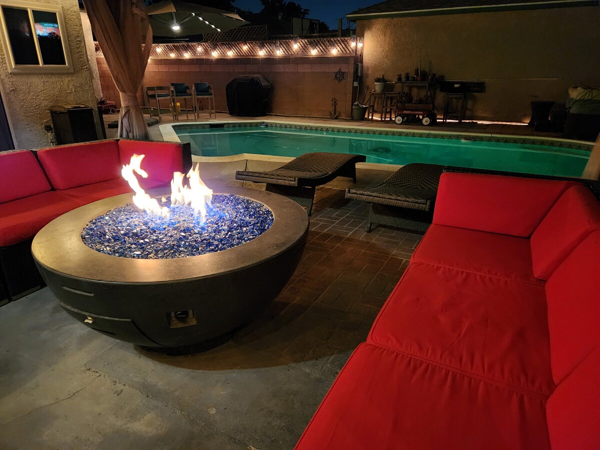 An outdoor seating area features a circular fire pit surrounded by red cushioned seating. Poolside lounge chairs are positioned near a well-lit swimming pool, with twinkling lights adorning the surrounding backdrop, creating a warm and inviting evening atmosphere.