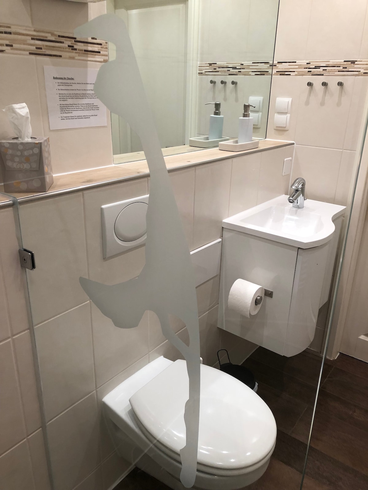 A modern bathroom is shown with a clear barrier featuring an abstract design. The space includes a compact toilet, a small sink, and various toiletries displayed on the counter. Light-colored tiles and a wooden floor contribute to a clean and functional environment.