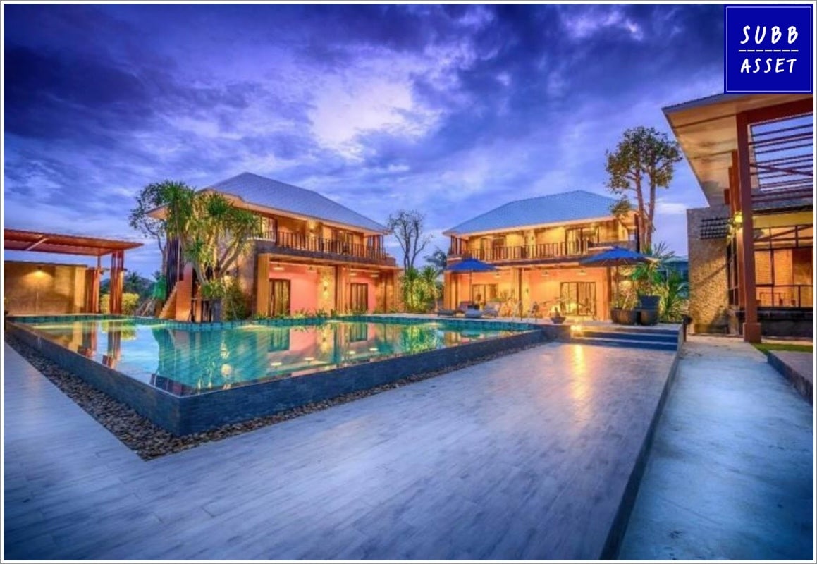 The image showcases a private pool villa featuring two adjacent buildings with expansive glass doors. A large swimming pool reflects the vibrant sky, surrounded by a wooden deck and lush greenery. The serene ambiance is enhanced by warm outdoor lighting and multiple shaded areas.