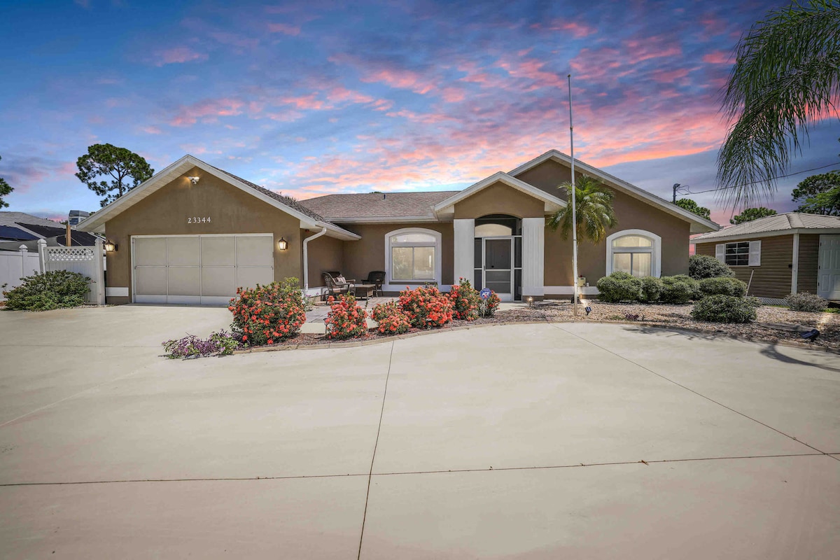 A large driveway leads to the entrance of the home, flanked by well-maintained landscaping with colorful flower beds. The exterior features a welcoming facade with large windows and a garage. The sky showcases vibrant hues of sunset, enhancing the home's inviting appearance.
