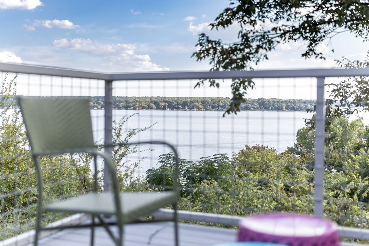 A private deck is shown overlooking Lake Worth, framed by lush greenery. A green chair is positioned prominently, offering a space to relax and enjoy the view of the calm water. The sky displays soft clouds, enhancing the serene atmosphere of the setting.