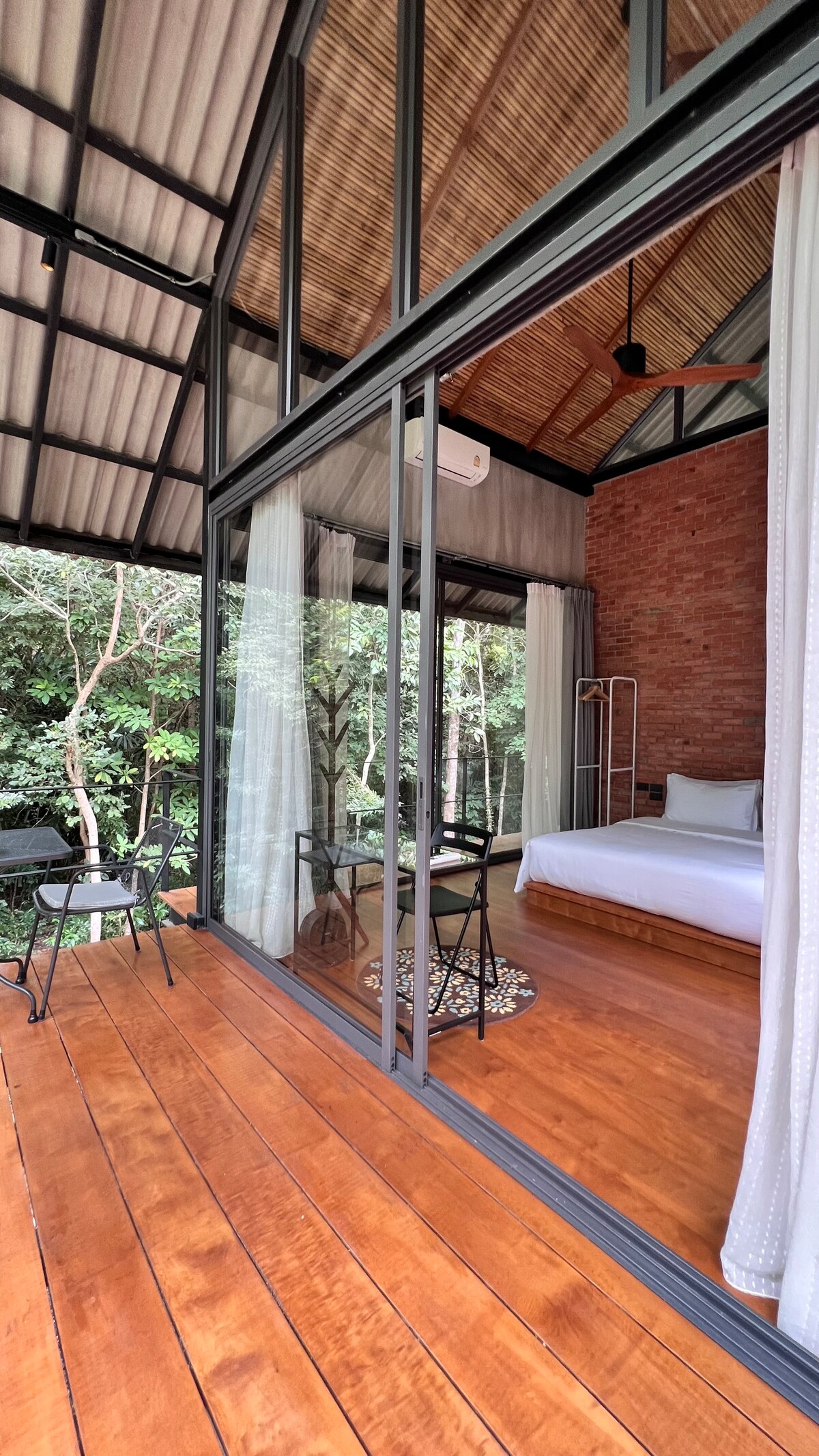 A spacious room is displayed through large glass doors that provide a view of the lush outdoor surroundings. Inside, a cozy bed is complemented by wooden accents, while a small table with two chairs sits on the wooden deck, inviting relaxation.
