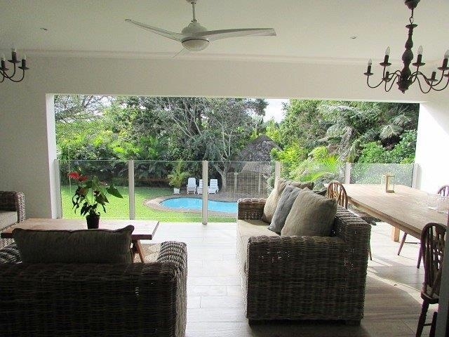An open living space features comfortable seating arrangements made of wicker. A large dining table is set beside a glass railing, providing a view of the lush garden and pool area just beyond. Sunlight filters in, creating a bright and refreshing ambiance.