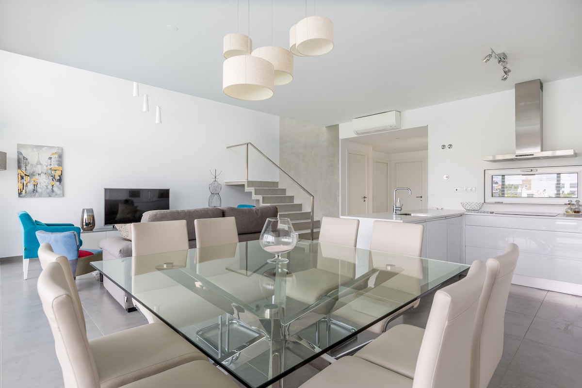 A modern dining area features a glass table surrounded by white leather chairs. Soft beige pendant lights hang above, illuminating the space. The open-plan design connects to a living area with a gray sofa and a television. A staircase leads to the upper level.