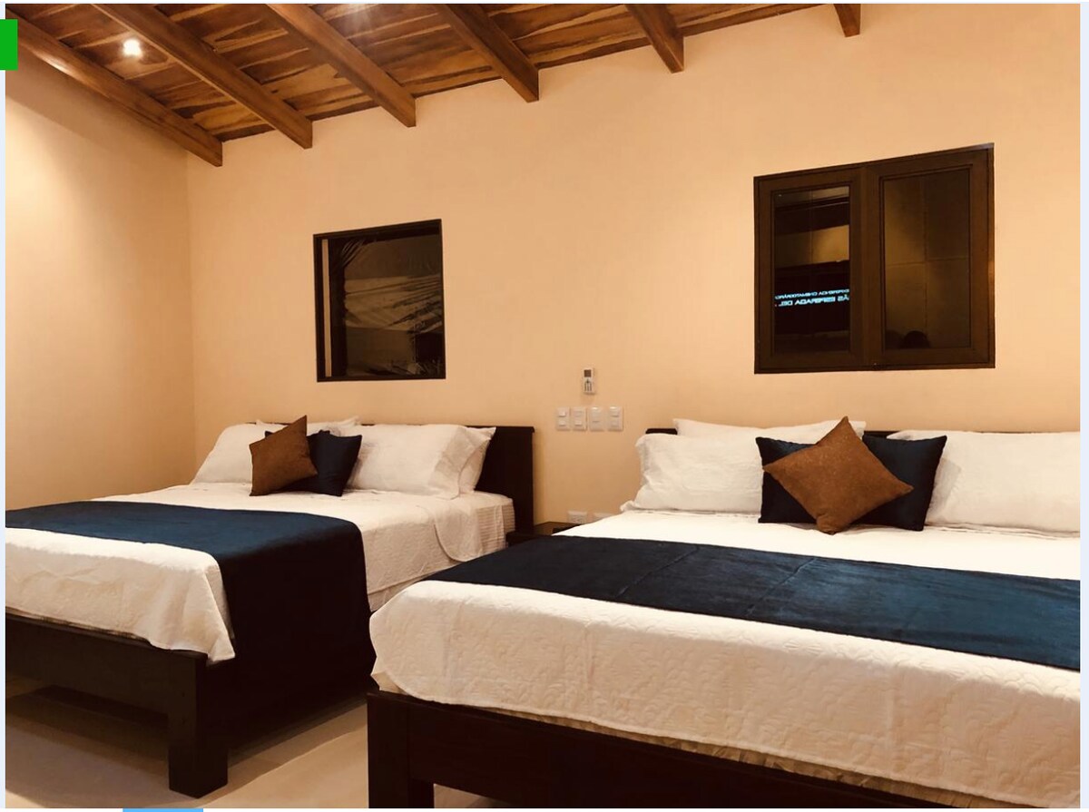 A suite features two spacious king beds, each adorned with neutral bedding and decorative pillows. The warm-toned walls complement the wooden ceiling, while two windows allow light to enter the room, enhancing the overall ambiance.