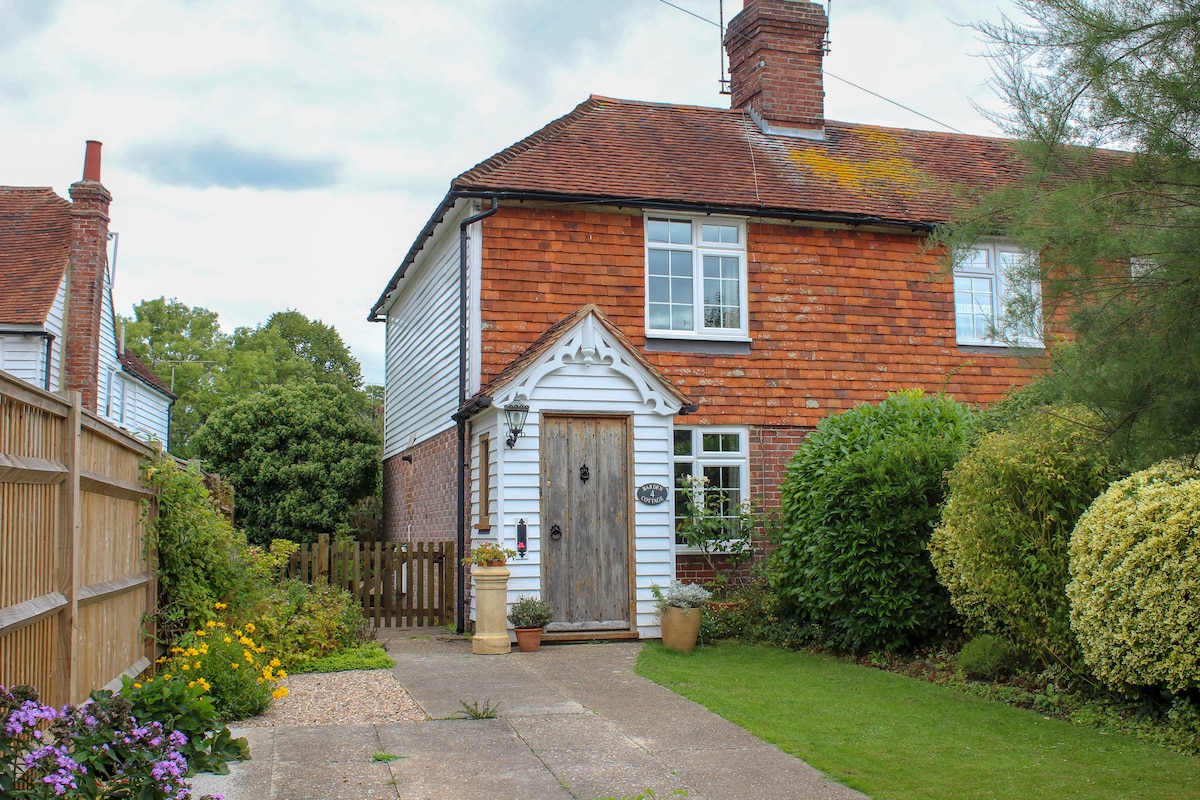 An inviting end terrace cottage with a mix of red brick and white siding is showcased. The entrance features a rustic wooden door and a charming pathway leading to it, bordered by colorful flowers and lush greenery. A well-maintained garden is visible on the side.
