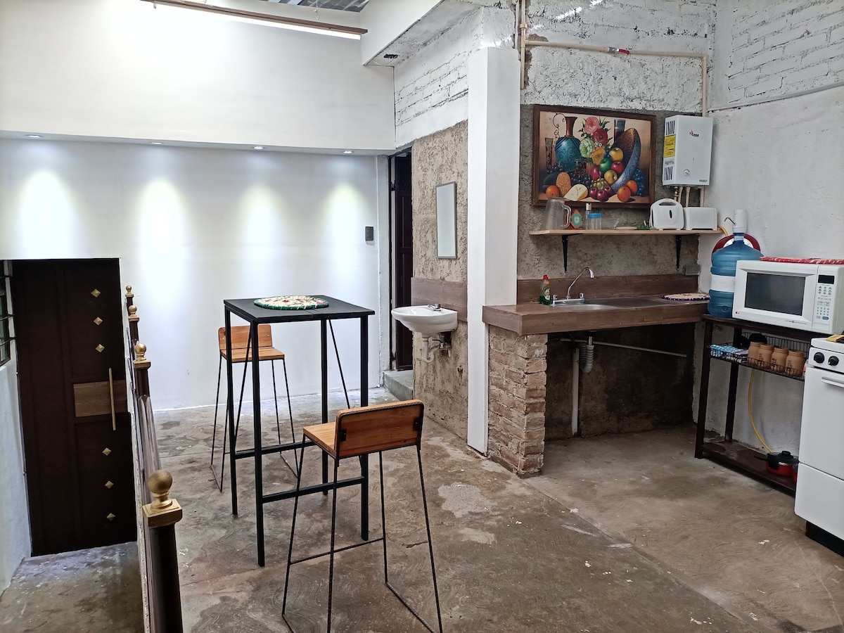 A compact kitchen area features a countertop with essential appliances, including a microwave and a stove. A dining table with two chairs provides a space for meals. The walls are accented with a colorful fruit painting, and the floor is finished with a simple concrete texture.