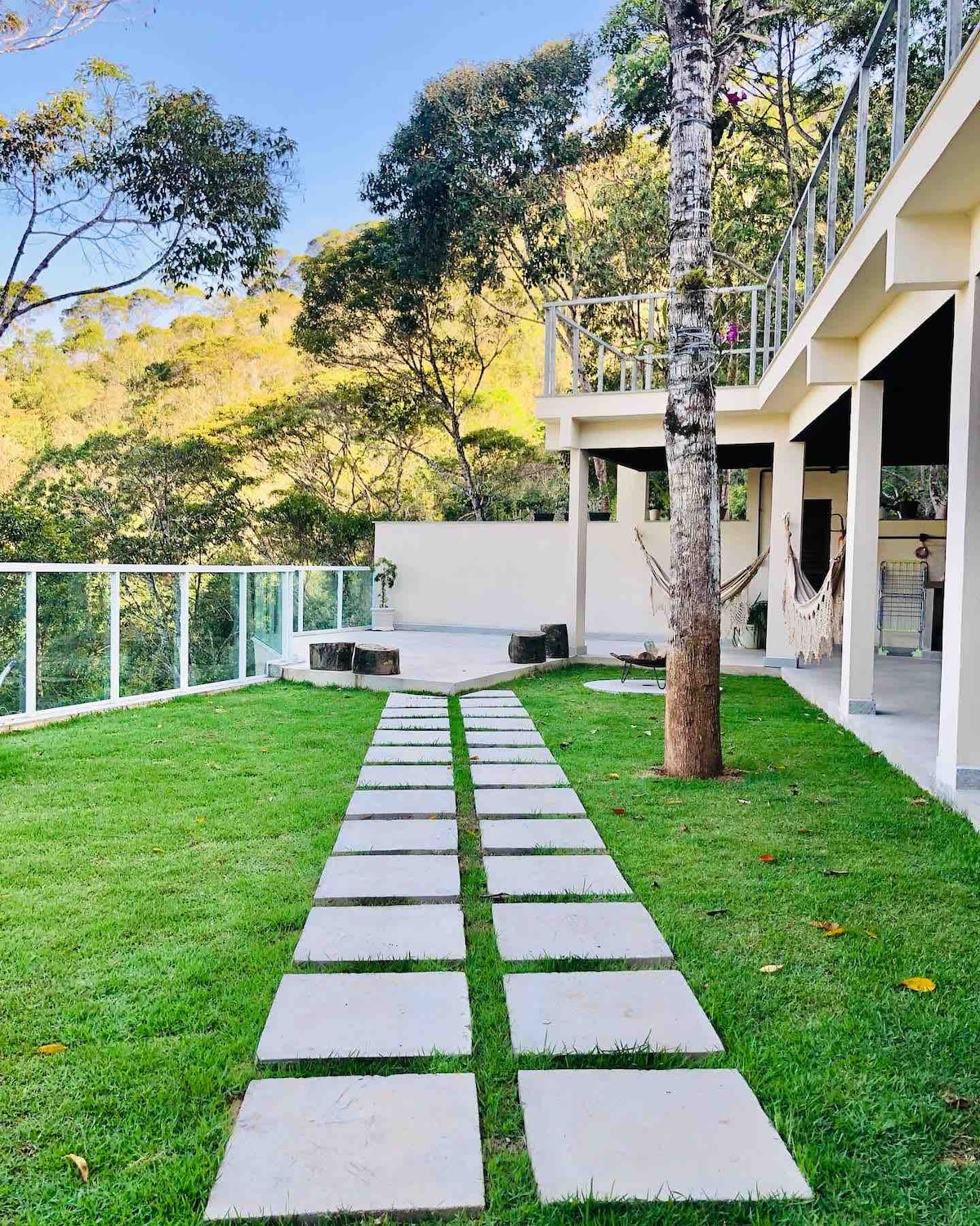 A well-maintained pathway of rectangular stone slabs runs across a green lawn, leading towards a modern house. The setting is surrounded by tall trees and overlooks a natural landscape, while a hammock hangs nearby, enhancing the outdoor space.