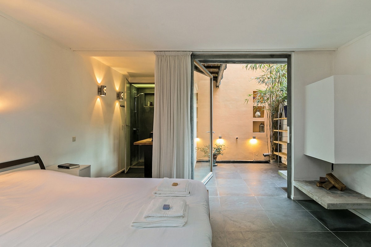 A spacious sleeping area features a neatly made bed with quality linens. Soft lighting illuminates the room, while large sliding doors provide a view into a tranquil courtyard filled with greenery. A small kitchenette and a stylish storage unit are also visible within the space.
