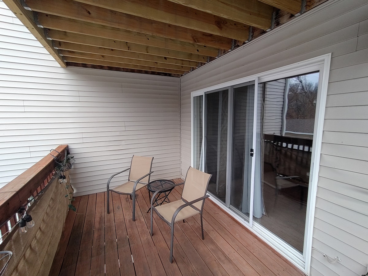 A private balcony area features two metal chairs and a small table on wooden flooring. The sliding glass doors allow natural light to enter, and a covered overhead provides protection from the elements. Simple railing offers a clear view of the surrounding area.