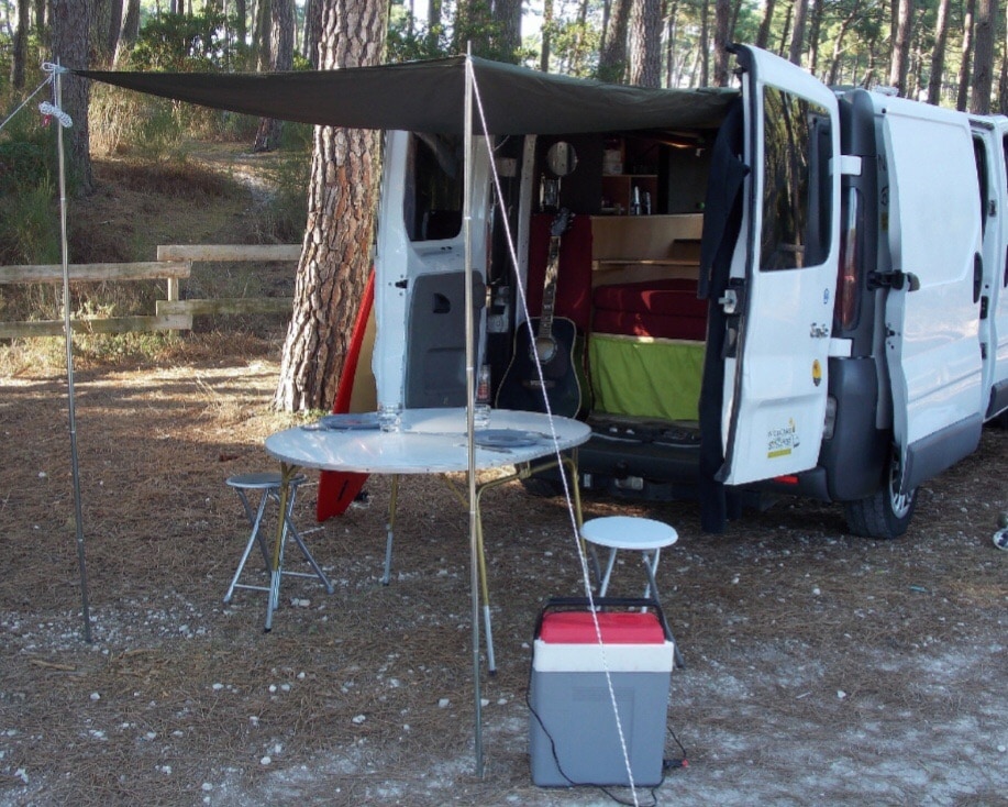 A parked van features an open side, revealing an organized interior with a bed and storage. A shaded area is set up outside with a round table and two chairs, alongside a cooler. Surrounding trees provide a natural backdrop to the setup.