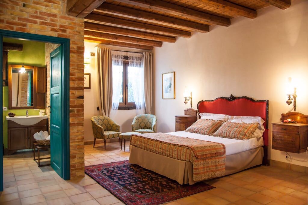 The spacious bedroom features a large bed with a patterned coverlet and a red headboard. Two armchairs are positioned near the window, and classic wooden chests of drawers complement the room. Exposed wooden beams and a terracotta floor enhance the rustic charm.