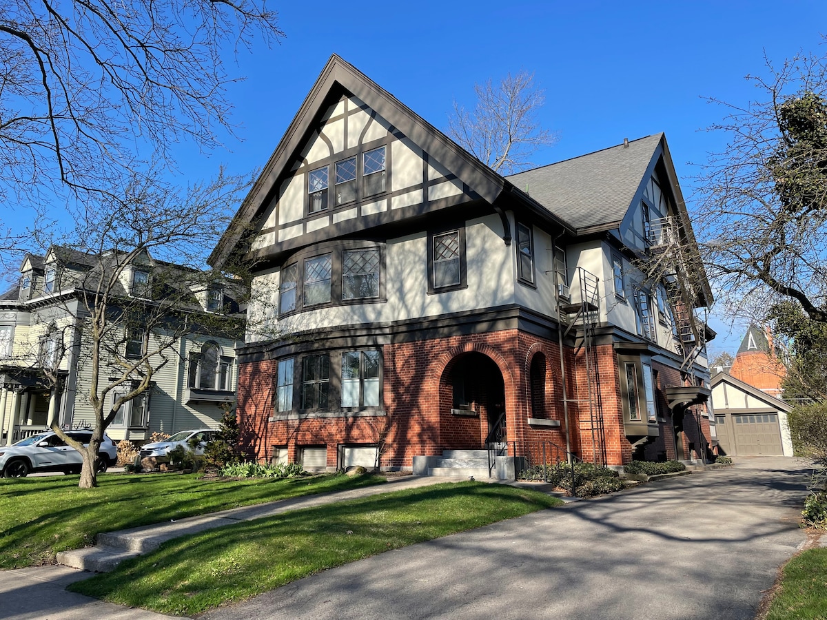A charming three-story building features a mix of brick and wood siding, highlighted by a steeply pitched roof. Green lawns and trees surround the entrance, with shadows cast on the path leading to the door. Clear blue skies create a serene backdrop.