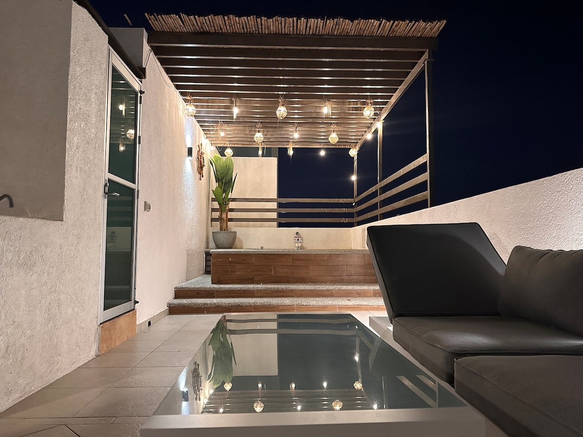 A rooftop area is illuminated by soft string lights, creating a relaxed ambiance. Comfortable seating is arranged around a glass-top table. A planter adds a touch of greenery, while a raised wooden platform provides additional seating or lounging space.
