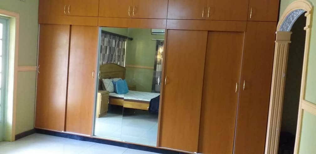 A spacious room is showcased with a large wooden wardrobe featuring mirrored doors. The reflection displays a bed with light bedding and decorative blue pillows. The walls are painted in a soft green, contributing to a calming environment.