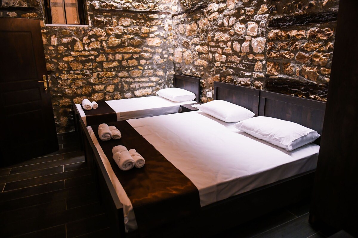 A room featuring two spacious beds with crisp white linens and four neatly rolled towels at the foot of each bed. The walls are composed of exposed stone, adding texture to the space. Soft lighting enhances the warm tones of the room.